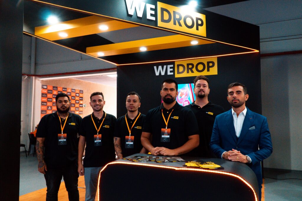 Na imagem, vemos seis homens em pé diante de um estande da WEDROP, uma plataforma de dropshipping on-line. Cinco deles vestem camisetas pretas com o logotipo da WEDROP e crachás laranja pendurados no pescoço. O Marcelo Menezes está à direita usando um terno azul. O estande é predominantemente preto com detalhes em amarelo e exibe o nome "WEDROP" em grandes letras brancas e amarelas no topo. O ambiente parece ser de uma feira ou conferência, sugerido pelo fundo decorado com logotipos e iluminação clara. Créditos da imagem à página WEDROP 2.0.